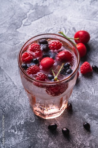 Fresh cold sparkling water drink with cherry, raspberry and currant berries in red faceted glass on stone concrete background, summer diet beverage, angle view