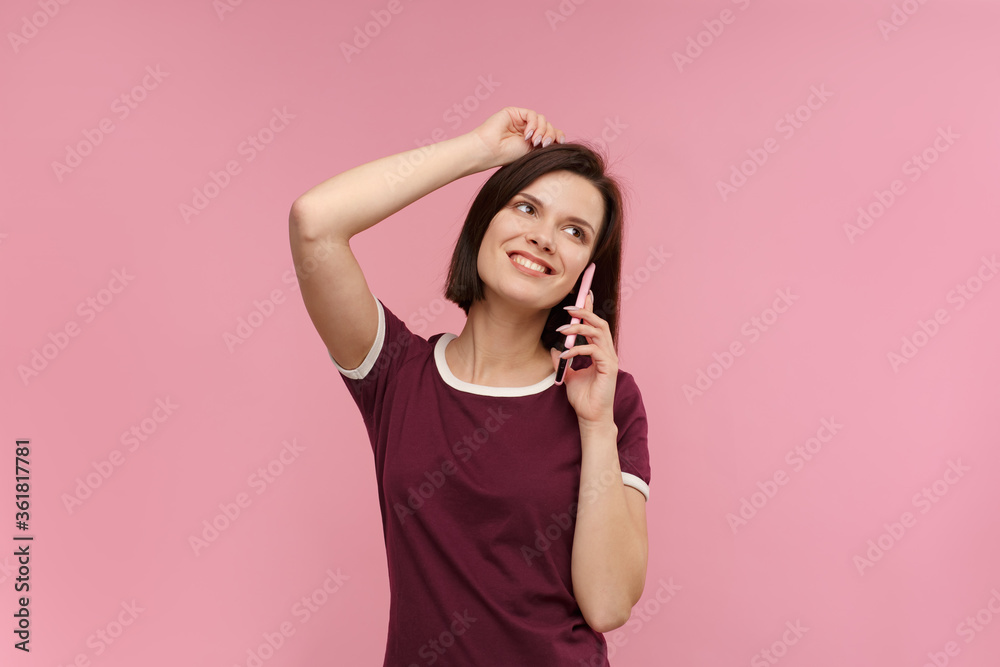 Fototapeta premium Young people working with mobile devices. Portrait of cute happy brunette woman wearing t-shirt talking on mobile phone and smiling isolated over pink background. Copy space.