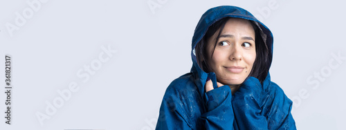 Portrait of a smiling girl dressed in blue raincoat in drops posing with hood on grey background in a studio.