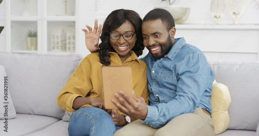 Young African American couple sitting on couch in living room and videochatting on tablet device. Male and female talking via webcam on computer at home on white sofa. Man and woman having videochat. Young African American couple sitting on couch in living room and videochatting on tablet device. Male and female talking via webcam on computer at home on white sofa. Man and woman having videochat.