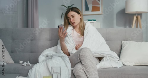 Ill young Caucasian woman with fever sitting on couch at home and videochatting on smartphone. Sick girl caughing and having videochat on mobile phone via webcam. Coronavirus isolation. photo