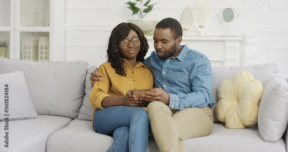 Young African American beautiful woman and handsome man sitting at home and talking. Couple having nice talk and using tablet device. Male and female at home hugging and chatting. Young African American beautiful woman and handsome man sitting at home and talking. Couple having nice talk and using tablet device. Male and female at home hugging and chatting.