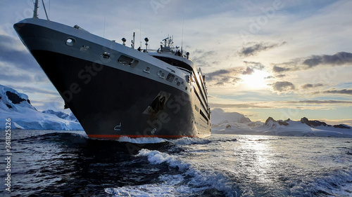 Cruise ship in Antarctic waters surrounded by glaciers and snow covered mountains with evening sun in the background 