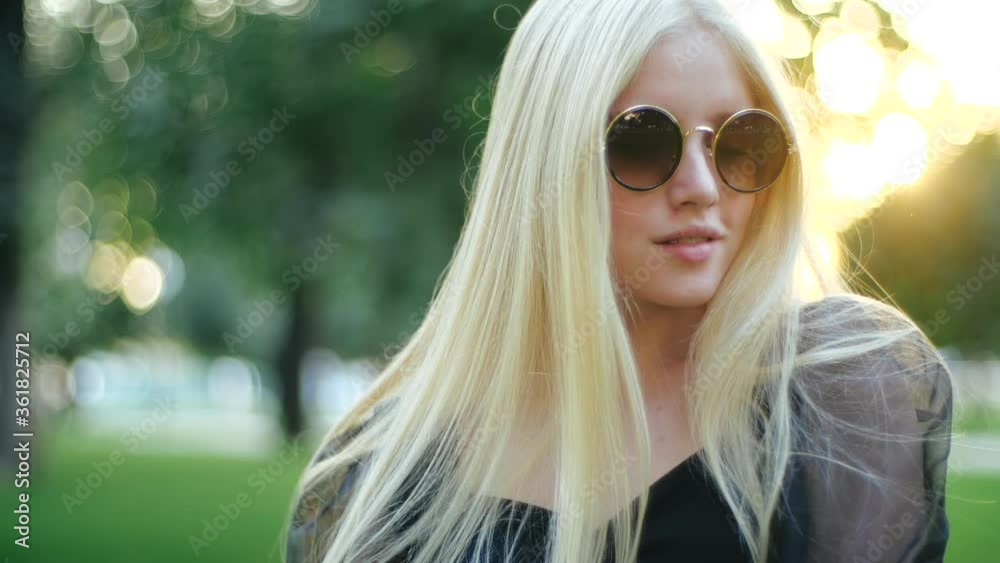 Stunning cool blonde with long hair in round sunglasses and light black dress beautifully poses for the camera in spring park against background of trees and grass. Photoshoot of young wonderful girl.