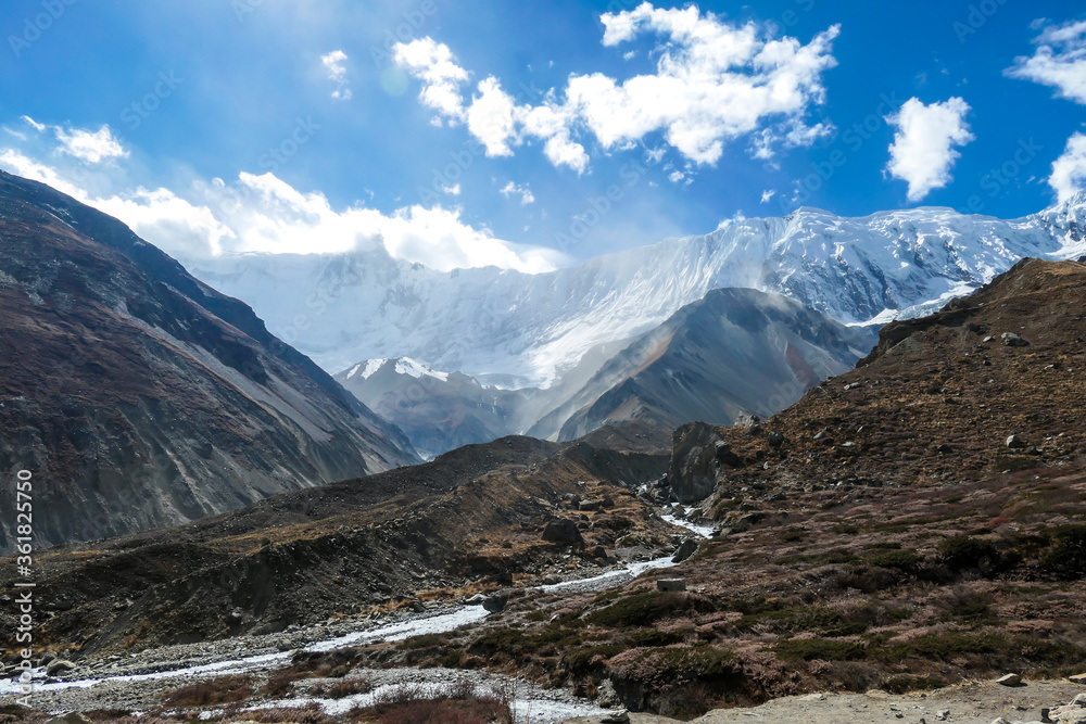 Panoramic view on a valley along Annapurna Circuit in Nepal, with a small torrent flowing in the middle. In the back there are high, snow capped Himalayan peaks. Slopes are overgrown with small bushes