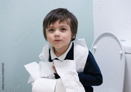 School kid playing with toilet paper in toilet,Healthy kid boy sitting with holding a roll of toilet paper and lookign at camera with smiling face,Child tear toilet roll against light green background photo