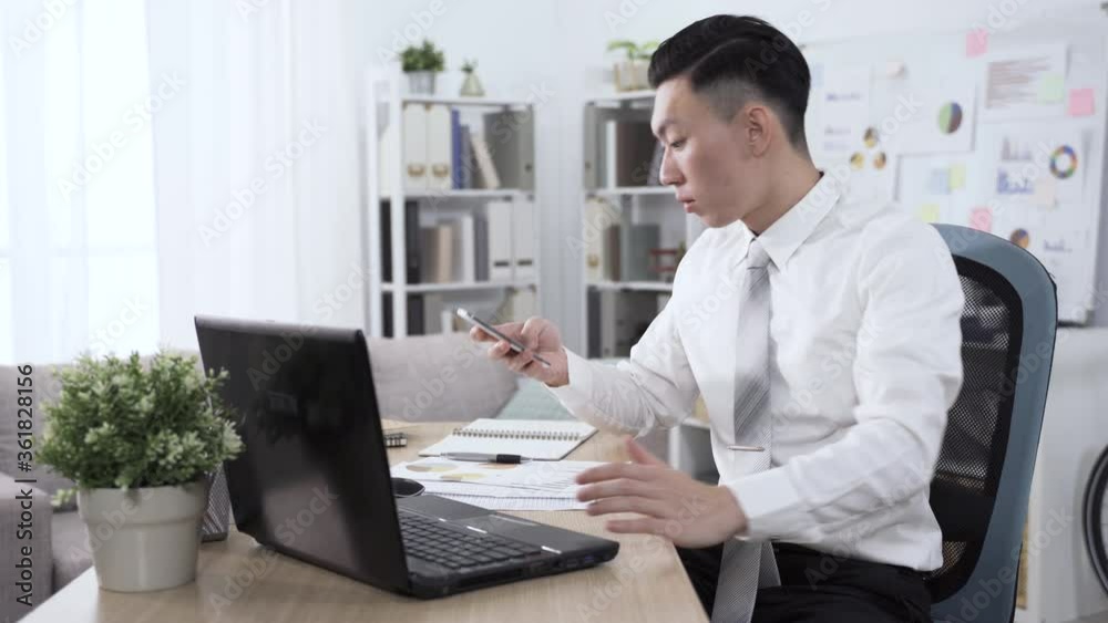 chinese guy sitting at desk typing is answering a phone. businessman leaning back comfortably in chair, putting hand in pocket and crossing legs while speaking on smartphone. chinese guy sitting at desk typing is answering a phone. businessman leaning back comfortably in chair, putting hand in pocket and crossing legs while speaking on smartphone.