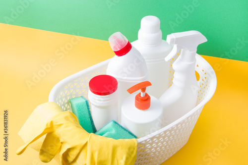 green sponges for washing, yellow rubber gloves, a yellow rag for cleaning and various white plastic bottles with detergents for the home in a basket on a yellow and green background
