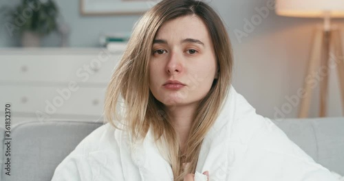 Portrait of young Caucasian bad-looking sick woman with coronavirus looking at camera and caughing in napkin while sitting on sofa in ivig room at home. Close up of ill girl. Self-isolated female. photo