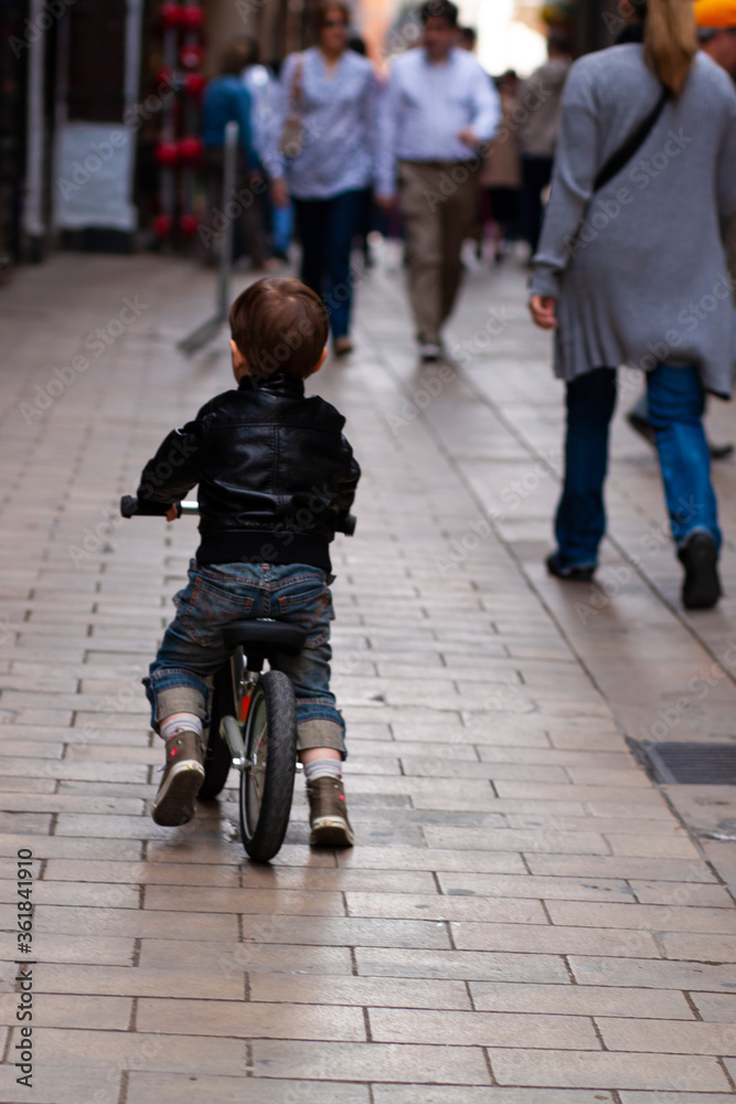 Close up image showing a cool little boy wearing stylish jeans and a leather jacket, riding a bike in a busy street. He is following his mom at a distance and is comfortable being in public .