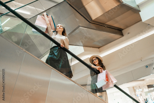 Two girls on an escalator in a shopping center. In the hands of paper bags. Shopping, leisure and entertainment.