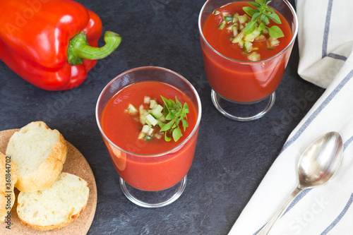 Summer diet. Cold tomato gazpacho soup in glass glasses with sliced cucumbers, peppers and greens on a black background top view. Sliced bread for snacks. Ice for cooling. Vegetarian food