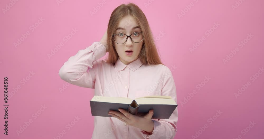 Charming young girl in eyewear reading book and looking at camera with shocked emotion. Blonde woman in pink shirt found some unexpected moment. Isolated over studio background. Charming young girl in eyewear reading book and looking at camera with shocked emotion. Blonde woman in pink shirt found some unexpected moment. Isolated over studio background.