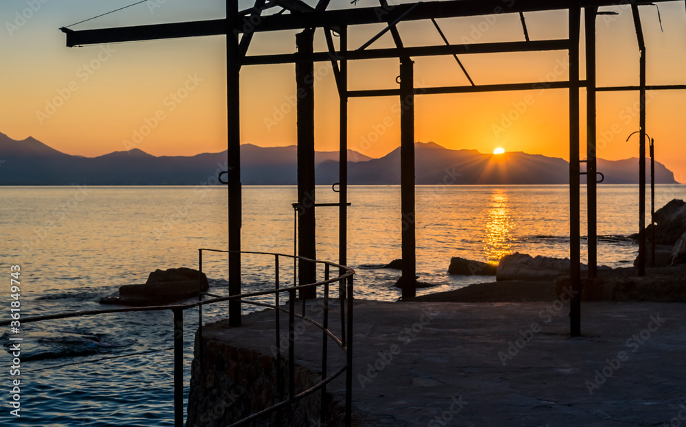 The sun dips below the mountains around in the Gulf of Palermo seen against the silhouette of a derelict boat hoist on a summer's evening
