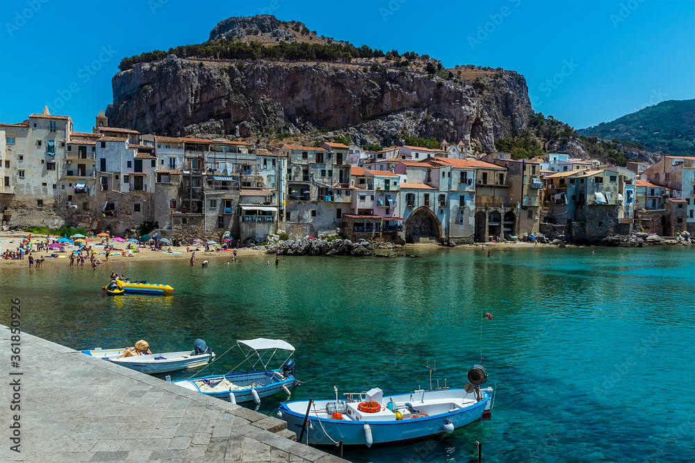 Small boats moored against the harbour walls of Cefalu, Sicily with the town and the Mesa as a backdrop it in summer