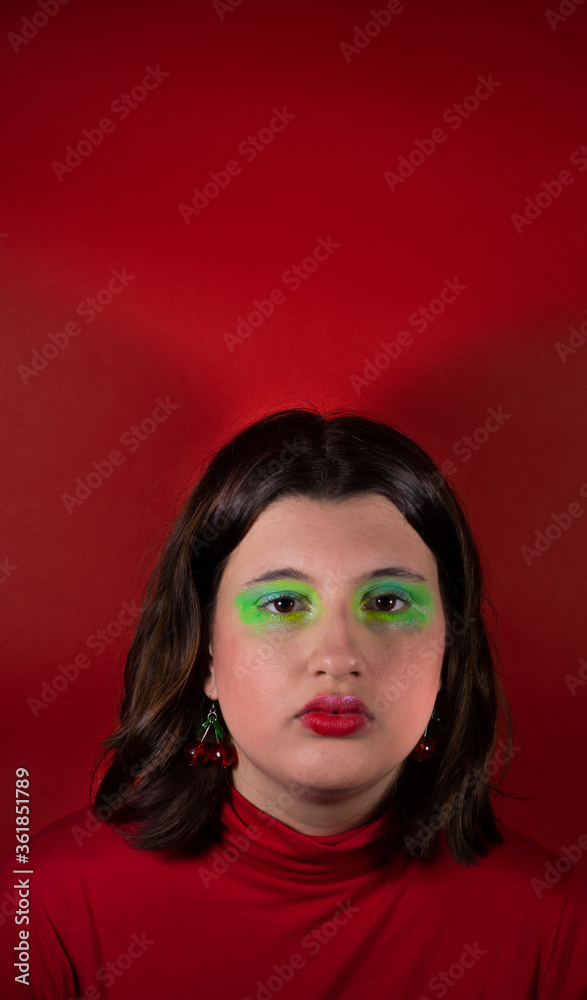 Modelo con maquillaje verde y pelo castaño mirando a cámara con fondo y remera roja Modelo con maquillaje verde y pelo castaño mirando a cámara con fondo y remera roja
