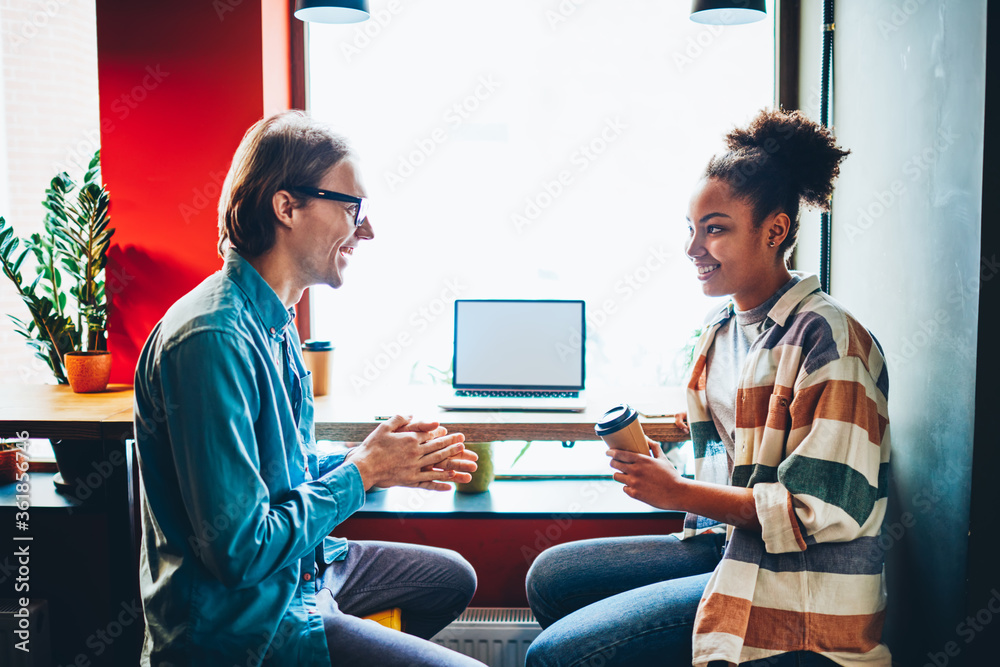 Cheerful multiracial male and female colleagues having conversation during coffee break,joyful students talking to each other and discussing plans sitting near laptop computer with blank screen.