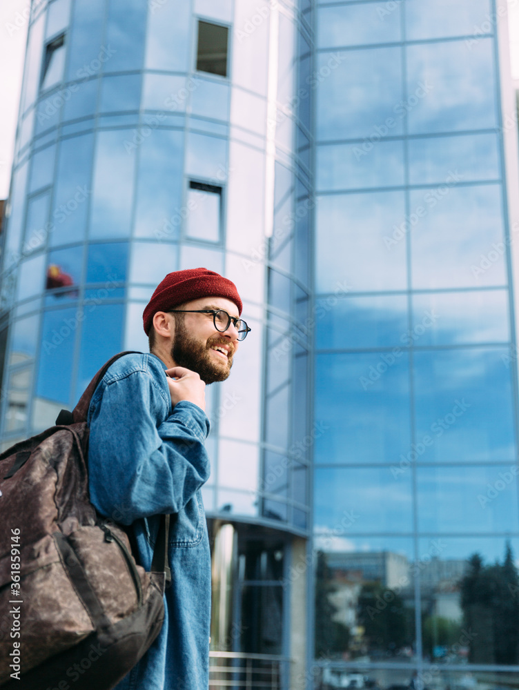 Handsome bearded male hipster student with backpack wearing round eyeglasses and red knitted hat looking away smiling