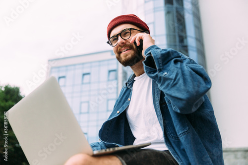 Closeup portrait of modern bearded male hipster in eyeglasses talking on phone and using laptop at the same time