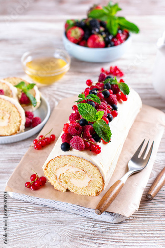 Sponge cake roll filling cream and berries, strawberry, raspberries, blueberry and red currants on white wooden background