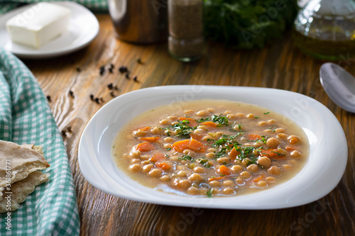 Vegetarian thick cheakpea soup with corrots and greens in white plate on wooden table with ingredients around. Turkish and greek cuisine. Healthy vegetable food. Horizontal. Etli Nohut, Revithia dish. photo