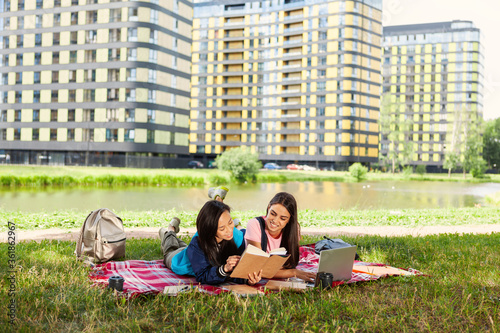 Two smiling female students doing homework together on lawn on campus after classes. Smiling girls lying on blanket, reading book and browsing laptop