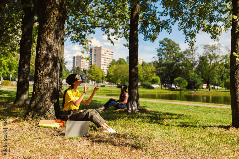 Side view of female student in VR headset swiping visualized data sitting under tree in park. Another college girl wearing virtual reality glasses in background Side view of female student in VR headset swiping visualized data sitting under tree in park. Another college girl wearing virtual reality glasses in background