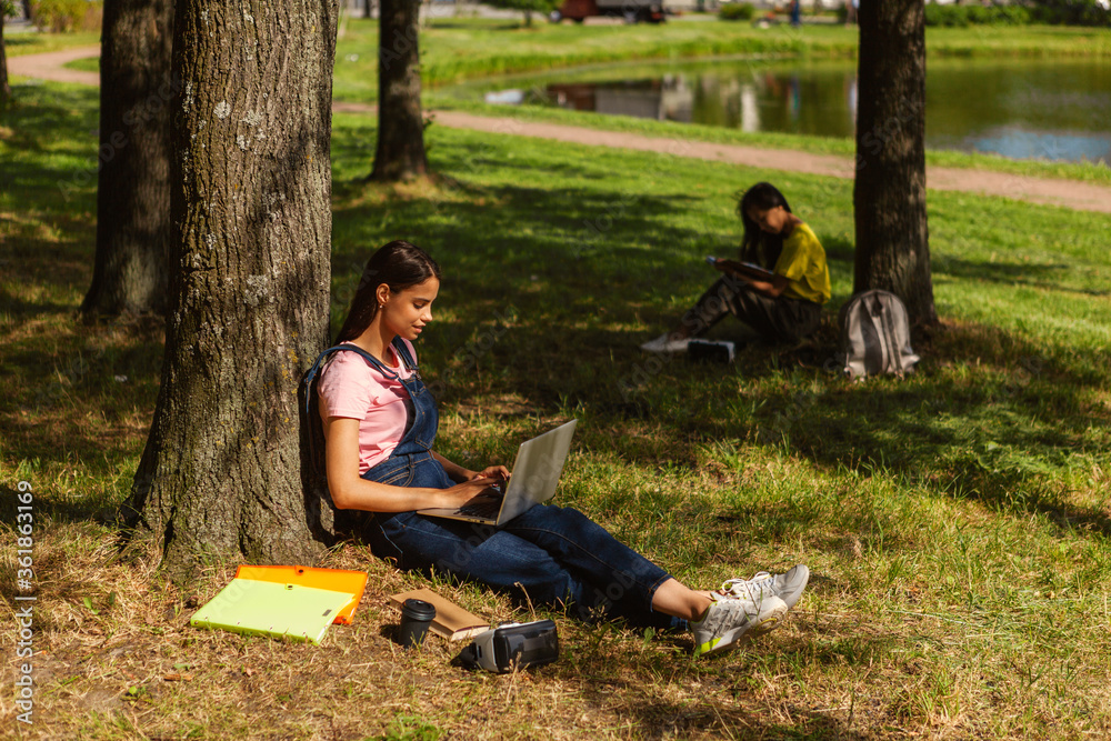 Side view of female student sitting under tree and typing on laptop in parkб textbooks, coffee cup and VR glasses nearby. Another college girl reading book in background