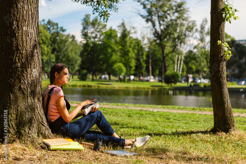 Side view of female student holding VR headset sitting under tree in park, looking away and thinking