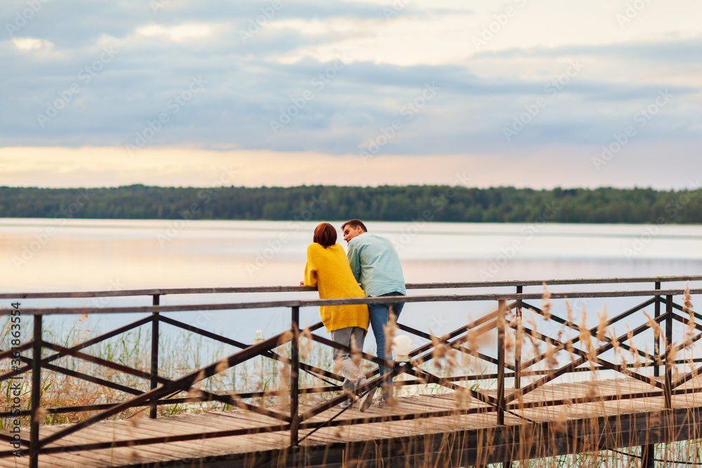 Rear view of romantic young couple standing on wooden pier leaning on railing, admiring beautiful river and sky scenery and talking