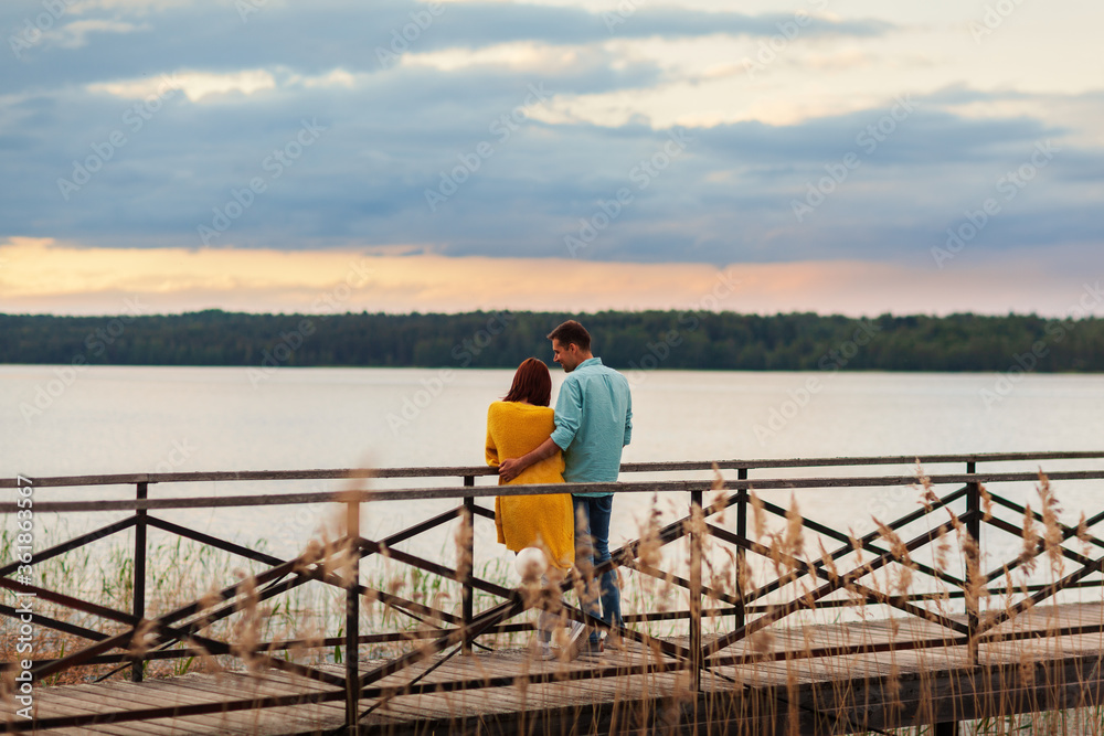 Back view of loving young couple embracing on wooden pier leaning on railing, admiring beautiful river view and talking cheerfully