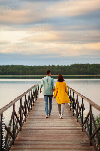 Rear perspective view of young romantic couple walking on wooden pier over river holding hands in summer evening