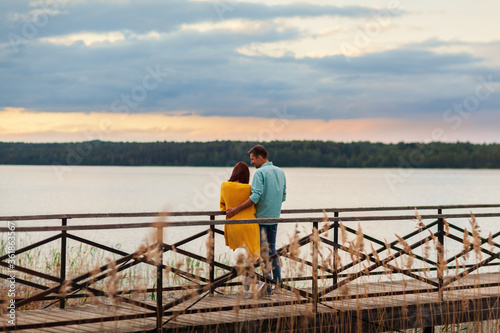 Back view of loving young couple embracing on wooden pier leaning on railing, admiring beautiful river view and talking cheerfully