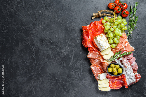 Italian antipasto, wooden cutting board with prosciutto, ham, parma, goat and Camembert cheese, olives, grapes. antipasti. Black background. Top view. Copy space