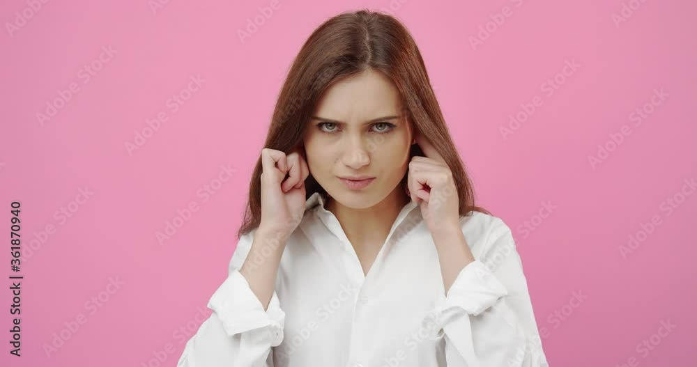 Annoyed young woman with dark hair plugging ears with hands while suffering from noise. Female model in white shirt avoiding loud sounds. Isolated over pink studio background. Annoyed young woman with dark hair plugging ears with hands while suffering from noise. Female model in white shirt avoiding loud sounds. Isolated over pink studio background.