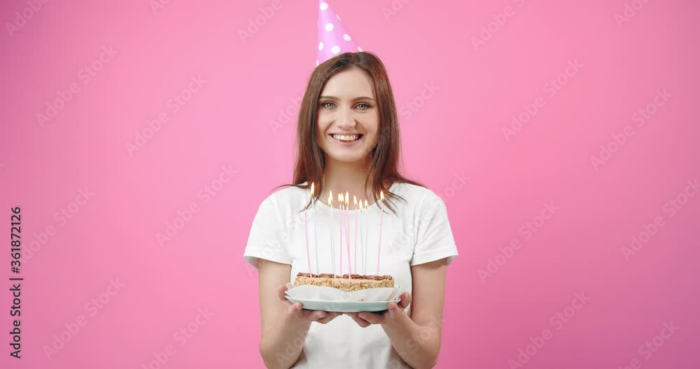 Cheerful brunette girl in white shirt and birthday cap holding delicious cake with burning candles while posing in studio with pink background. Concept of celebration and happiness. Cheerful brunette girl in white shirt and birthday cap holding delicious cake with burning candles while posing in studio with pink background. Concept of celebration and happiness.