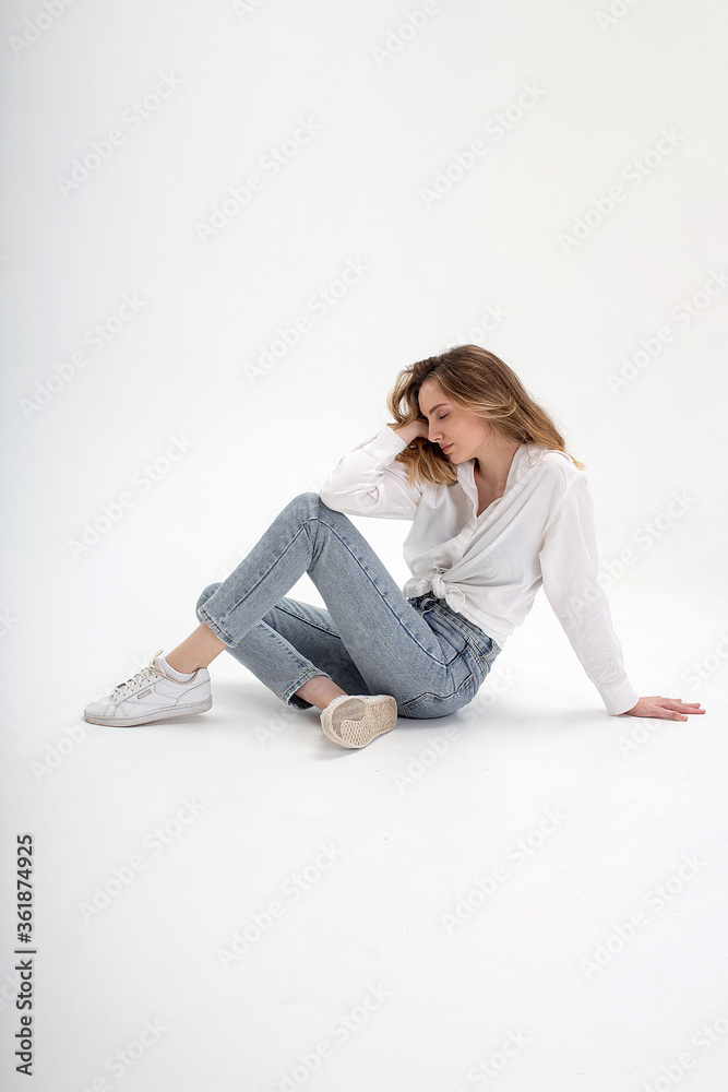 portrait of young caucasian woman posing in shirt and blue jeans, sitting on white studio floor. model tests of pretty girl in basic clothes on cyclorama. attractive female poses with eyes closed portrait of young caucasian woman posing in shirt and blue jeans, sitting on white studio floor. model tests of pretty girl in basic clothes on cyclorama. attractive female poses with eyes closed