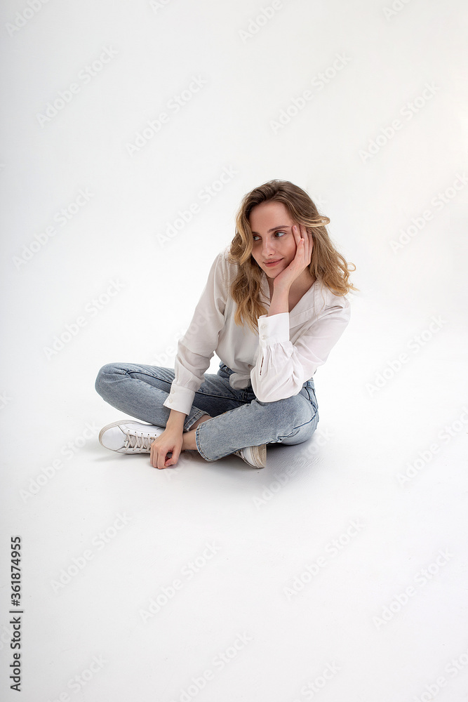 portrait of young smiling caucasian woman posing in shirt and blue jeans, sitting on white studio floor. model tests of pretty girl in basic clothes on cyclorama. attractive female poses, smiles