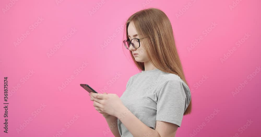 Side view of concentrated young girl in eyeglasses using smartphone for sending text messages. Beautiful woman with blond standing in studio over pink background. Side view of concentrated young girl in eyeglasses using smartphone for sending text messages. Beautiful woman with blond standing in studio over pink background.