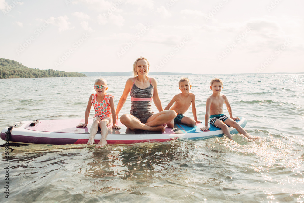 Obraz premium Caucasian woman parent sitting on paddle sup surfboard in water with kids children. Modern outdoors family activity. Individual summer aquatic recreation sport hobby. Healthy lifestyle.