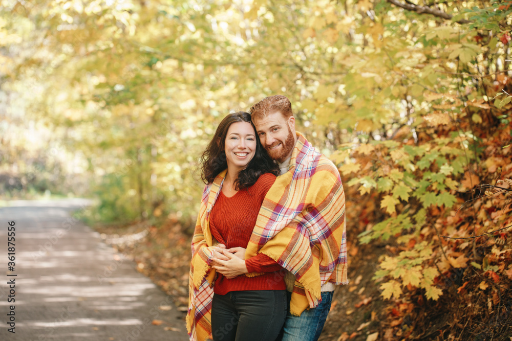 Fototapeta premium Beautiful couple man woman in love. Boyfriend and girlfriend wrapped in yellow blanket plaid hugging together in a park on autumn fall day. Tenderness and happiness. Authentic real people.