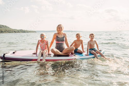 Caucasian woman parent sitting on paddle sup surfboard in water with kids children. Modern outdoors family activity. Individual summer aquatic recreation sport hobby. Healthy lifestyle.