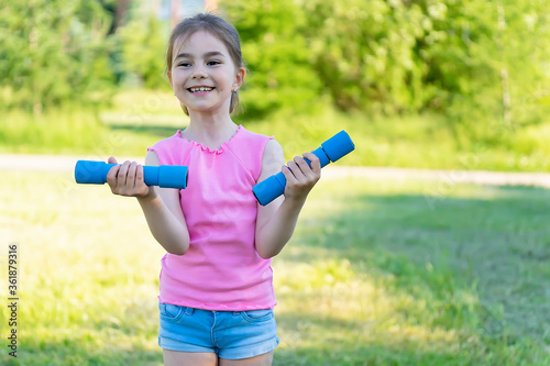 The child goes in for sports in the park. Little girl is engaged with dumbbells.