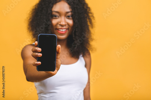 Portrait of a smiling young african american black woman holding blank screen mobile phone isolated over yellow background.