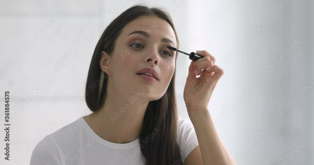 Confident happy young woman holding mascara apply on eyeleashes prepare getting ready in the morning, smiling beautiful girl doing make up put cosmetics on face looking in mirror in bathroom at home