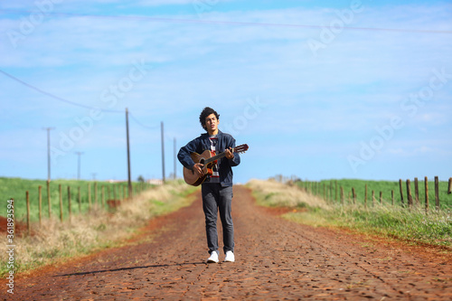 Jovem afroamericano posa tocando violão em estrada rural em dia ensolarado com céu azul e nuvens de fundo