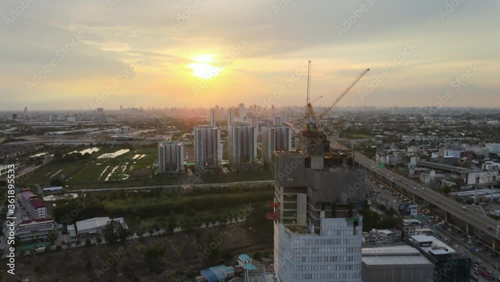 Aerial Shot of the Skyscraper Building in the Process of Construction. In the Background Working Crane, Forest, sea and Sunset. Shot on Aerial Shot of the Skyscraper Building in the Process of Construction. In the Background Working Crane, Forest, sea and Sunset. Shot on