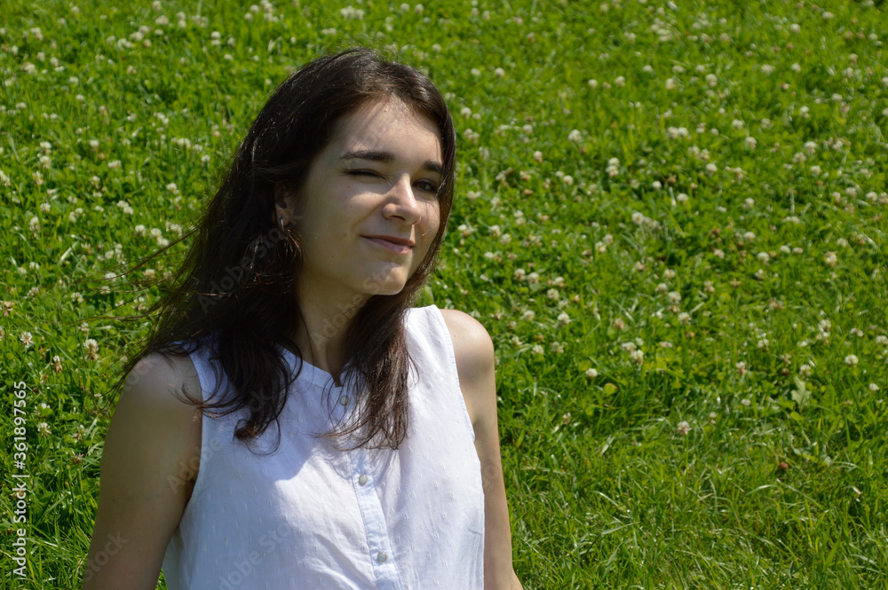 Portrait of young brunette woman winking and looking in the camera on a background of green meadow with clover Portrait of young brunette woman winking and looking in the camera on a background of green meadow with clover
