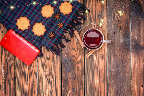 Christmas tartan plaid scarf, fairy lights, gingerbread biscuits, cup of red karkade tea and cinnamon sticks on dark rustic wooden background. Christmas flat lay with space for text