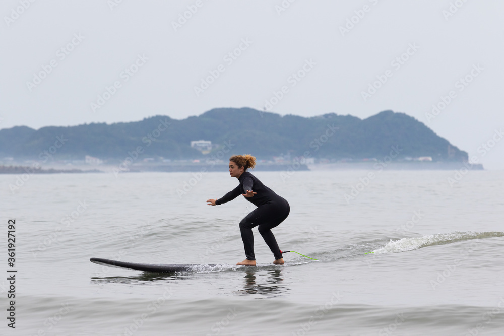 Young multiracial African American lady with amazing smile, freckles & frizzy hair, learning to surf wearing a black wetsuit on a black surfboard. She is Surfing in Chiba Japan near to Ichinomiya. Young multiracial African American lady with amazing smile, freckles & frizzy hair, learning to surf wearing a black wetsuit on a black surfboard. She is Surfing in Chiba Japan near to Ichinomiya.